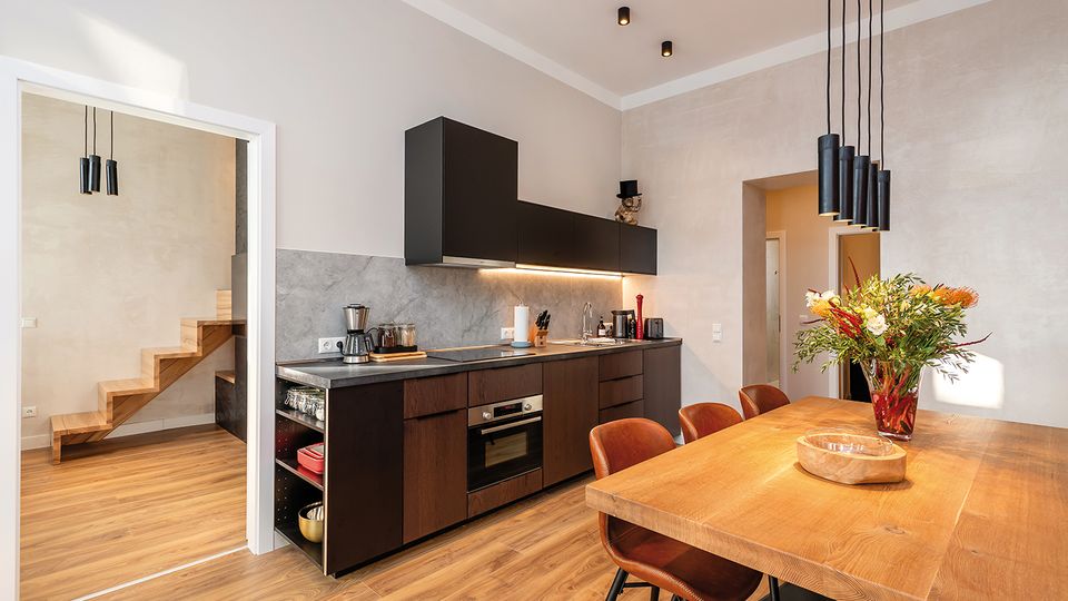 Moderne Wohnküche mit dunklen Hängeschränken und Holzfronten, grauer Arbeitsplatte und durchgehendem Fliesenspiegel sowie integrierten Geräten. Im Vordergrund steht ein großer Holztisch mit Stühlen unter schwarzen Pendelleuchten; links führt ein offener Durchgang zu einer Holztreppe.