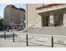 Straßenszene mit einem größeren Gebäudeeingang aus Stein mit breiten Treppen und Blumen auf dem Balkon rechts. Links im Bild stehen ein gelbes Wohnhaus mit Geschäften im Erdgeschoss und ein Eingang zur U-Bahn-Station mit Schild "U" und Passanten im Vordergrund.