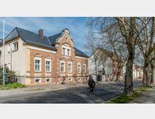 Straßenszene mit einem historischen Wohngebäude aus gelblich-braunem Backstein und weißen Fenstereinfassungen, das sich auf der linken Seite befindet. Im Vordergrund fahren ein Radfahrer und eine Person läuft auf einem Gehweg entlang; rechts sind Bäume mit kahlen Ästen zu sehen.