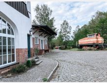 Rustikaler Hof mit gepflastertem Vorplatz, links ein Gebäude mit weiß verputzter Wand, Ziegelsteinfundament und großen Fenstern, rechts eine orangefarbene, alte Lokomotive in einer grünen, von Bäumen umgebenen Umgebung.