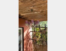 Ein überdachter Außenbereich mit einer Decke aus Bambus oder Schilfrohr, an der ein Kronleuchter mit lilafarbenen Lampenschirmen hängt. Im Hintergrund ist eine rosafarbene Wand, ein Fensterrahmen und eine Holzrankhilfe mit Pflanzen zu sehen.