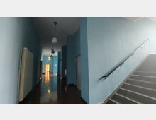 Ein Flur in einem Gebäude mit hellblauen Wänden, mehreren weißen Türen und einer Treppe mit schwarzem Handlauf auf der rechten Seite. Von der Decke hängen runde Hängeleuchten, und der Boden ist glänzend.