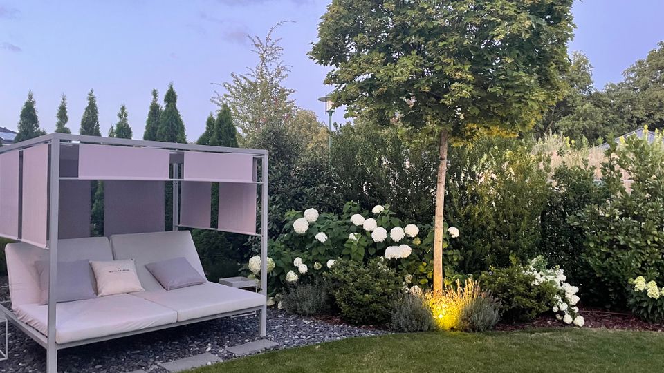 Moderner Gartenbereich mit Rasenfläche, einem weißen Daybed mit Rahmen und Sonnensegel auf Kiesbett sowie dichtem Grün aus Hecken und Sträuchern. Rechts steht ein kugelförmig geschnittener Baum mit Bodenbeleuchtung, umgeben von weißen Hortensienblüten.