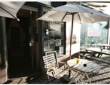 Außenbereich eines kleinen Cafés mit runden Holztischen und Stühlen unter großen Sonnenschirmen auf Kopfsteinpflaster. Dahinter stehen eine Glasfront mit Tür und Fenster sowie eine offene Eingangstür in den Innenraum.