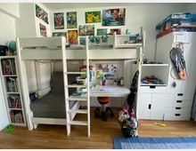 Kinderzimmer mit Hochbett und integriertem Schreibtisch. An der Wand sind bunte Poster, daneben stehen ein Bücherregal, weiße Staumöbel sowie ein Basketballkorb; auf dem Fußboden liegt ein Holzboden mit einem blauen Teppich.