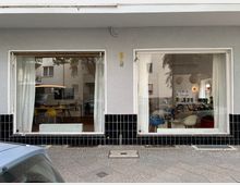 Fassade eines Gebäudes mit zwei großen Schaufenstern, durch die ein Innenraum mit modernen Möbeln sichtbar ist, darunter gelbe und weiße Stühle sowie Tische. Der untere Bereich der Fassade ist mit schwarzen Fliesen verkleidet, darüber eine helle Putzwand, und im Vordergrund ist ein Gehweg zu sehen.