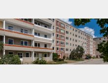 Wohnblock aus der Nachkriegszeit mit fünf Stockwerken, grauen Fassaden und weißen Fenstern. Im Vordergrund sind kleine Grünflächen, Büsche und Gehwege zu sehen, bei sonnigem Wetter mit blauer Himmel und wenigen Wolken.