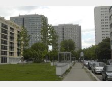Stadtlandschaft mit großen Wohnhochhäusern, Grünflächen und einer Gehwegpromenade am rechten Bildrand. Im Vordergrund Bäume und ein moderner gläserner Wartebereich, daneben geparkte Autos entlang der Straße.