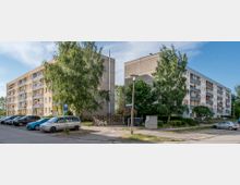 Zwei mehrstöckige, rechteckige Wohnblöcke in einem Wohngebiet, umgeben von Bäumen und geparkten Autos. Die Gebäude haben Balkone und eine sandfarbene sowie graue Fassade.