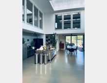 Ein modernes, lichtdurchflutetes Loft mit einer offenen Küche aus Edelstahl, hohen Decken und einem großen Oberlicht. Im Hintergrund befindet sich ein Essbereich mit Tisch und Stühlen, vor einer großen Glasfront mit Blick ins Freie.