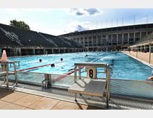 Freibad in einem Stadion mit rechteckigem Schwimmbecken, umgeben von Tribünen aus Stein und einem historischen Gebäudekomplex im Hintergrund. Im Becken schwimmen Menschen, und im Vordergrund ist ein Startblock mit der Nummer 8 sichtbar.