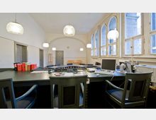 Heller Konferenzraum mit hohen, gewölbten Fenstern, mehreren Schreibtischen und Stühlen sowie Computern und Büchern auf den Tischen. Im Hintergrund befinden sich Türen, eine Garderobe und eine Bank, beleuchtet von mehreren Hängelampen.