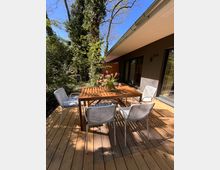 Eine Terrasse mit Holzboden, darauf ein rechteckiger Holztisch mit vier weißen Stühlen und einem Blumenstrauß in der Mitte. Im Hintergrund sind eine Hausfassade mit Glasfenstern und ein dicht bewachsener Garten mit Bäumen zu sehen.