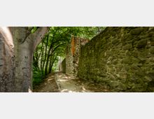 Ein schmaler, von Bäumen gesäumter Weg verläuft entlang einer historischen Steinmauer, die aus unregelmäßig angeordneten Natursteinen und einigen Backsteinen besteht. Das dichte Blätterdach der Bäume wirft Schatten auf den Weg und die Mauer.