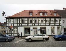 Fachwerkhaus mit weißer und dunkelgrüner Fassadengestaltung, roten Dachziegeln und Schaufenstern im Erdgeschoss, gelegen an einer gepflasterten Straßenecke mit parkenden Autos und Verkehrsschildern. Im Vordergrund sind ein Stoppschild, Fußgänger und Fahrzeuge zu sehen.