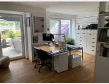 Helles, modernes Arbeitszimmer mit einem Schreibtisch aus Metall und Holz, darauf ein Computer und Schreibtischlampe. Im Raum befinden sich weiße Regale und Schubladenschränke sowie große Fenster mit Blick auf eine Terrasse mit Gartenmöbeln und Pflanzen.