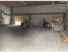 Eine leere Lagerhalle mit Betonboden, weißen Backsteinwänden und einer Holzdecke. Im Hintergrund befinden sich Fenster mit abgedecktem Glas, gestapelte Baumaterialien, eine Hebebühne sowie vereinzelte Möbel und Werkzeuge.