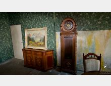 Ein Raum mit einer gemusterten Tapete und teils abgeblätterten Wänden. Zu sehen sind ein antiker Schrank mit Intarsien, darauf ein gerahmtes Gemälde einer Berglandschaft, eine hohe Standuhr aus Holz und ein Stuhl mit gepolsterter Rückenlehne.