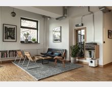 Moderner Wohnraum mit hellen Wänden und Holzboden, eingerichtet mit einer schwarzen Ledercouch, Holzstühlen und einem niedrigen Couchtisch auf einem grauen Teppich. An der Wand befinden sich ein großes Fenster, Bilder, Regale mit Büchern und Pflanzen, sowie eine Holzkommode mit Schallplatten und Musikanlage.