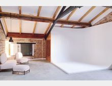 Industrie-inspirierter Innenraum mit Backsteinwänden, freiliegenden Holzbalken und einer weißen Fotostudio-Hohlkehle. Im Vordergrund stehen zwei hell gepolsterte Sessel mit passendem Hocker auf einem Betonboden.