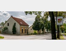 Straßenszene in einer ländlichen Gegend mit einem zweistöckigen Wohnhaus mit gelber und weißer Fassade und rotem Ziegeldach, umgeben von Bäumen. Rechts steht ein Schild mit der Aufschrift „Willkommen im Landkreis Potsdam-Mittelmark“.