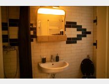 Badezimmer mit cremefarbenen Fliesen und schwarzen Akzenten, einem beleuchteten Spiegel über einem Waschbecken mit Wasserhahn und einem schwarzen Toilettensitz. Rechts ist ein Handtuchheizkörper und links eine Dusche mit Duschvorhang sichtbar.