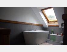 Modernes Badezimmer mit weißer freistehender Badewanne unter einer schrägen Decke. Ein Dachfenster mit Holzrahmen lässt Tageslicht herein, daneben steht ein kleiner Schrank mit grünen Türen und Dekorationen.