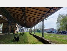 Ansicht unter einem großen, gewölbten Dach mit Holzverkleidung und Stahlstreben, das von Metallpfeilern gestützt wird. Im Hintergrund sind ein Industriegebäude, parkende Fahrzeuge, Bäume und ein grasbewachsener Bereich zu sehen.