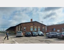 Backsteinbau mit Satteldach, flankiert von einem Parkplatz mit mehreren geparkten Autos. Im Vordergrund eine Person auf einem gepflasterten Bereich, bei bewölktem Himmel.