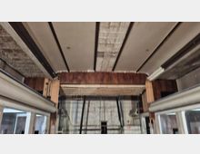 Blick nach oben in einen Industrie- bzw. Werkstattraum mit breiten Deckenbalken, großen Raster-Leuchtpanelen und freigelegten Holz- und Metallkonstruktionen. Seitlich sind Fensterfronten und Stahlträger sichtbar, im Hintergrund eine geflieste Wand mit Kabeln und Installationen.