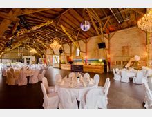 Geräumiger Veranstaltungsraum in einem rustikalen Backsteingebäude mit holzsichtbarem Dachstuhl, dekoriert mit Lichterketten, Kronleuchtern und weißen, gedeckten Tischen. Im Hintergrund befindet sich eine Theke aus Holz sowie helle Fenster und eine Tür.