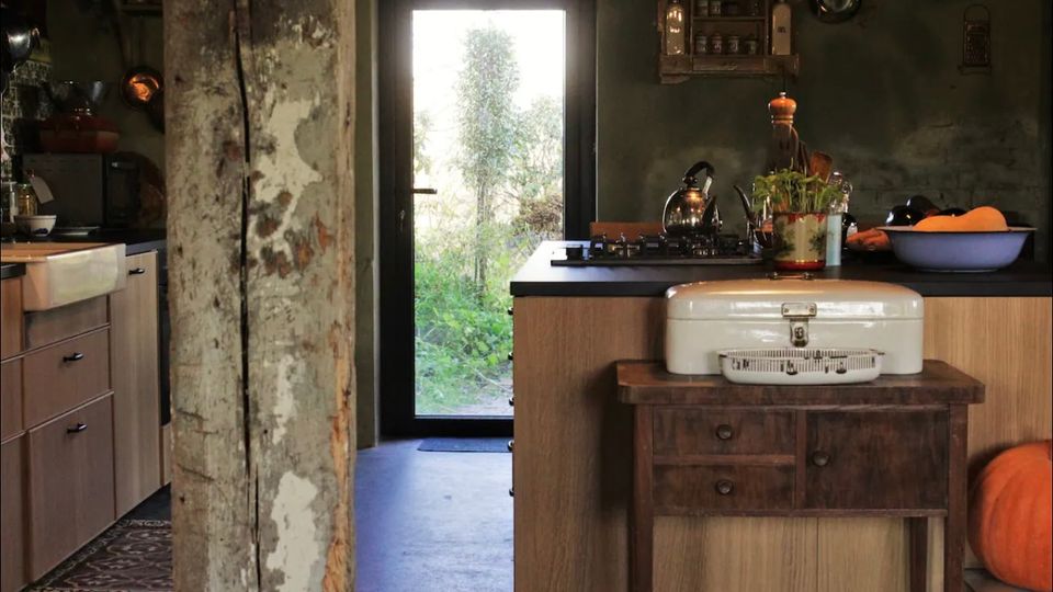 Rustikale Küche mit Holzmöbeln, einer freistehenden Kücheninsel und einer schwarzen Glastür, die nach draußen in einen grünen Garten führt. Eine alte Holzsäule teilt den Raum, Wände und Dekorationen wirken im Vintage-Stil.