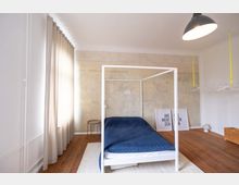 Minimalistisch eingerichtetes Schlafzimmer mit einem weißen Himmelbett und blauer Decke, Holzboden und einer Wand in Betonoptik. Links hängen helle Vorhänge, rechts ist eine offene Garderobenstange erkennbar, und an der Wand stehen zwei gerahmte Poster.