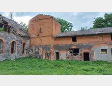 Ehemaliges Gebäude aus roten Backsteinen und Bruchsteinen mit beschädigtem Dach und zerbrochenen Fenstern, umgeben von einer Wiese. Die verfallene Struktur verfügt über mehrere Bögen und steht in einer grünen Umgebung mit Bäumen im Hintergrund.