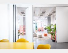 Heller, moderner Büroraum mit großen Glas-Schiebetüren, hohen Decken mit Akustikpaneelen und viel Tageslicht. Im Hintergrund stehen orange Regale und Tische, mehrere Pflanzen sowie runde Sitzhocker auf einem Teppich; vorne ist ein gelber Tisch mit Stühlen zu sehen.
