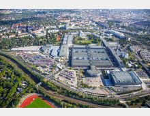 Luftaufnahme eines großen Messegeländes mit mehreren rechteckigen Hallen und angrenzenden Parkplätzen, umgeben von Bahngleisen, Straßen, Parks und Wohngebieten. Im Vordergrund befinden sich ein Sportplatz mit roter Tartanbahn und Grünflächen sowie dichte Vegetation.