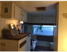 Innenansicht eines Wohnwagens mit schlichter Einrichtung. Links ein Holzarbeitsbereich mit Spiegel und Deko wie einer Pflanze, im Hintergrund ein kleines Sofa vor einem großen Fenster mit Blick auf eine Wand außerhalb.