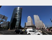 Straßenkreuzung am Potsdamer Platz in Berlin mit mehreren modernen Hochhäusern, darunter ein abgerundetes Glasgebäude und ein rechteckiger, heller Büroturm. Im Vordergrund fahren Autos vorbei, während Menschen auf der Straße unterwegs sind.