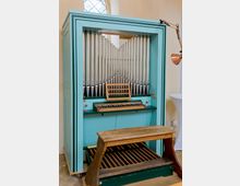 Eine kleine, türkisfarbene Pfeifenorgel in einem Innnenraum mit hellem Wandton. Sichtbar sind die Pfeifen, eine Klaviatur, ein Pedalbrett und eine Holzbank; im Hintergrund ein schlichtes, bogenförmiges Fenster.