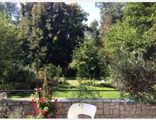 Garten mit dichter grüner Vegetation, großen Bäumen und gepflegtem Rasen, im Vordergrund eine Natursteinmauer mit Blumen und ein weißer Gartenstuhl. Im Hintergrund ist ein kleiner Holzbogen und eine Gartenliege erkennbar.