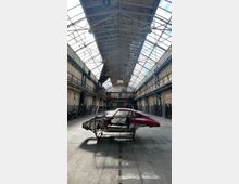 Große Industriehalle mit hohem Glasdach und sichtbaren Stahlträgern, flankiert von gemauerten Seitenwänden mit Rohrleitungen und Laufstegen. In der Mitte steht ein teilweise zerlegter Autokarosserie-Rohbau auf einem Metallgestell auf dem Betonboden.