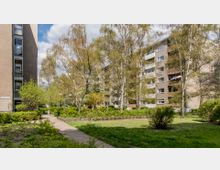 Wohnanlage mit mehreren beige-braunen Mehrfamilienhäusern und Balkonen, umgeben von einer Grünanlage mit Rasen, Bäumen, Sträuchern und einem gepflasterten Gehweg. Im Hintergrund sind helle Wolken und blauer Himmel sichtbar.