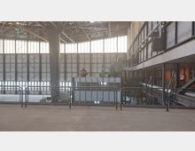 Industriehalle mit hoher Decke, großen Fensterfronten aus Glas und Stahlrahmen, und massiven Stahlträgern. Im Vordergrund ist ein Metallgeländer, dahinter technische Anlagen und eine erhöhte Galerie mit Treppen und Plattformen sichtbar.