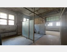 Ein leerstehender, verfallener Raum mit rohem Ziegelmauerwerk, mehreren Fenstern mit Metallrahmen und Lichtzufuhr, sowie gefliesten Wänden und Böden. In der Mitte befindet sich eine gläserne Trennwand, von der alte Rohrleitungen und Leitungen ausgehen.