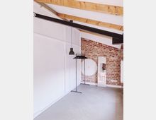 Ein heller Raum mit sichtbaren Holzbalken an der Decke, einer weißen und einer Ziegelwand. Der Raum enthält eine schwarze Hängelampe, zwei Kleiderstangen, einen hohen Spiegel und ein rundes Fenster in der Ziegelwand.