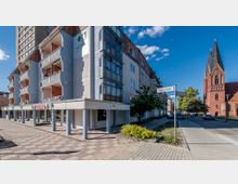 Wohngebäude mit Balkonen und einem überdachten Eingangsbereich an einer Straßenecke, daneben eine rote Backsteinkirche mit hohem Turm. Im Vordergrund sind Straßenschilder und ein Gehweg mit Bäumen im Hintergrund zu sehen.