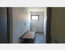 Ein kleiner, karger Raum mit weißen Betonwänden, einem kleinen Fenster oben an der Rückwand, einem einfachen Holzbettgestell an der linken Wand und einem Heizkörper unter dem Fenster. Der Boden ist aus grauem Beton, und die Tür zum Raum steht offen.