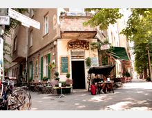 Ecke eines historischen Gebäudes mit abgenutzter Fassade, in dem sich das Café und Kneipe „Herman Schulz“ befindet. Vor dem Eingang stehen Sitzgelegenheiten, grüne Fensterläden, eine handgeschriebene Tafel und Pflanzen, während Fahrräder und ein ruhiger Gehweg im Schatten der Bäume zu sehen sind.