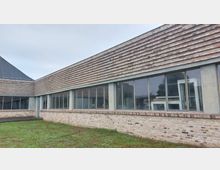 Flaches Gebäude mit moderner Architektur, bestehend aus einer Fassade aus Ziegelstein und großen Fensterfronten mit schmalen Rahmen. Im Vordergrund befindet sich eine Rasenfläche, während der Himmel wolkenverhangen ist.