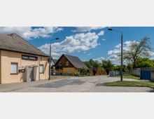 Straßenszene in einer ländlichen Umgebung mit einem beige gestrichenen Gebäude auf der linken Seite, beschriftet mit „Fränkis Tanzbar“, daneben ein modernes gelbes Einfamilienhaus mit dunklem Satteldach, umgeben von Gärten und Bäumen. Der Himmel ist blau mit vereinzelten Wolken, und im Vordergrund sind Straßenlaternen und asphaltierte Wege zu sehen.